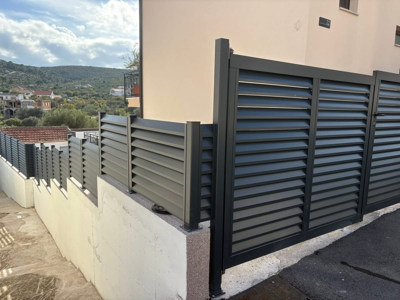 Antracitna aluminijska ograda s kosim lamelama i dvokrilna vrata iste izvedbe uz ulazni prilaz obiteljske kuće, s pogledom na zeleno brdo.